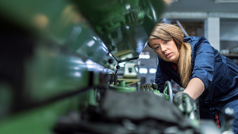 Photo of young brown hair woman in blue uniform wearing Protective Gloves at metallurgy factory. Beauty professional factory female employee using industrial machine to milling components. Beautiful Caucasian woman as industrial worker at metal sheet profiling machine at manufacturing factory.