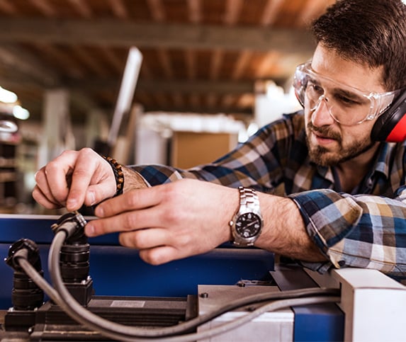 White middle-aged manufacturing professional wears safety glasses and ear protection while performing maintenance on a machining part