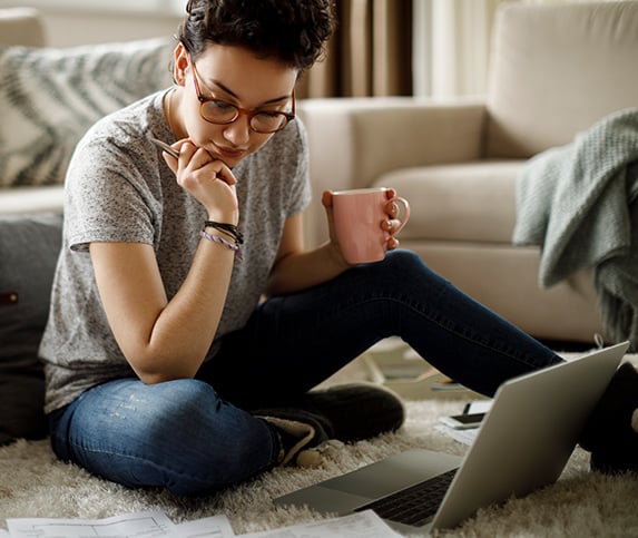 Young female workforce professional sits on a carpeted floor in front of a laptop holding a pen and a pink coffee mug while taking online Tooling U-SME classes