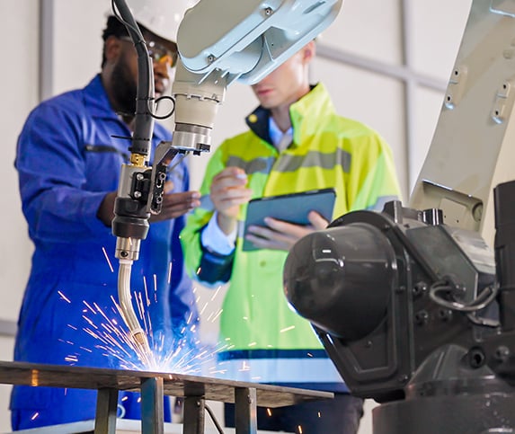 Two manufacturing professionals work on a tablet in a smart factory while a robotic arm welds together two metal plates